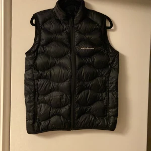 Svart väst  från Peak Performance - Säljer en svart väst från Peak Performance i storlek 160. Västen har en dragkedja framtill och är perfekt för kyliga dagar. Den är i nyskick och har en stilren design som passar både till vardags och utomhusaktiviteter. Perfekt för höst och vinter!