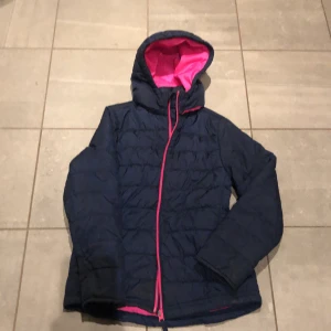 Mörkblå pufferjacka från True North - Säljer en snygg mörkblå pufferjacka från True North med en cool rosa insida. Jackan har en dragkedja i rosa och en praktisk huva. Perfekt för kyliga dagar och håller dig varm och bekväm. Passar bra för både höst och vinter!