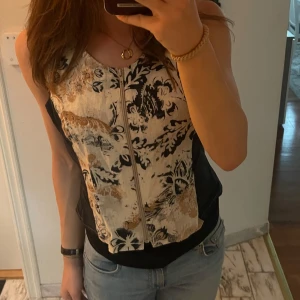 Mönstrad korsett med dragkedja - Snygg väst med ett unikt mönster i marinblå och beige. Den har en dragkedja framtill och är ärmlös, vilket gör den perfekt för sommardagar eller en utekväll. Passar bra med jeans eller kjol för en chic look. Säljer då den är lite stor. Står inre storlek men skulle säga M/L