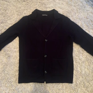 Svart cardigan med knappar - Säljer en stilren svart cardigan med knappar framtill. Perfekt för både vardag och fest. Den har en klassisk krage och två fickor framtill. Den är köpt i Rom, Italien så är väldigt exklusiv.