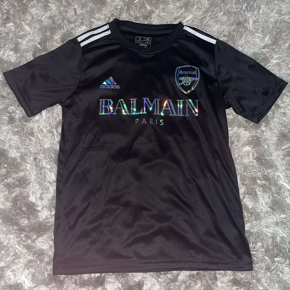 Säljer en snygg svart Arsenal fotbollströja från Adidas. Tröjan har korta ärmar och ett coolt Balmain Paris-tryck på framsidan. Den är i nyskick och kommer i originalförpackning. Perfekt för alla Arsenal-fans!. T-shirts.