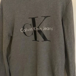 Grå tröja från Calvin Klein Jeans - Säljer en snygg och stilren grå tröja från Calvin Klein Jeans. Den har ett stort CK-logotyptryck på framsidan och är perfekt för en casual look. Tröjan är långärmad och gjord i ett mjukt material som är superbekvämt att ha på sig. Perfekt för höst och vinter!