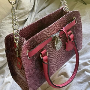 Rosa axelväska med ormskinnsmönster - Säljer en supercool rosa MK väska med ormskinnsmönster. Den har en snygg kedja som axelrem och detaljer i silver. Väskan är rymlig och perfekt för både vardag och fest. Den har en dragkedja för säker förvaring. 