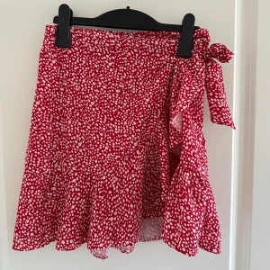 Röd prickig omlottkjol - Säljer en superfin röd omlottkjol med vita prickar. Kjolen har en söt knytning i sidan och är perfekt för sommardagar. Materialet känns lätt och luftigt, vilket gör den idealisk för varma dagar. Den har en volangdetalj som ger en extra touch av stil!