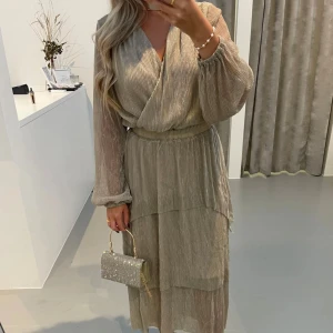 Beige långklänning med volangdetaljer - Säljer en elegant beige långklänning med volangdetaljer. Klänningen har en omlott-design och långa ärmar, vilket ger en stilren och sofistikerad look. Perfekt för festliga tillfällen! Passar även M.