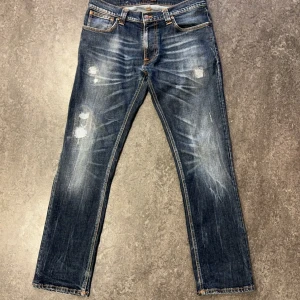 Blå jeans med slitningar - Säljer ett par Nudie Jeans med slitningar och en vintage look. De har en klassisk femficksdesign och orange sömmar som ger en schysst kontrast. Perfekta för en avslappnad stil!
