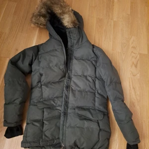 Grön pufferjacka med fuskpäls - Säljer en grön pufferjacka från Everest. Den är perfekt för vintern med sin varma vaddering och fuskpäls på huvan. Jackan har en dragkedja framtill och praktiska fickor. Den är i bra skick och redo för kalla dagar!
