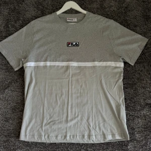 Grå t-shirt från FILA - Säljer en snygg grå t-shirt från FILA. Den har en klassisk design med en vit rand över bröstet och FILA-loggan broderad ovanför. Perfekt för en avslappnad stil, oavsett om du ska till gymmet eller bara hänga med kompisar. T-shirten är i bomull och har korta ärmar.