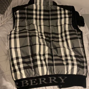 Svart och rutig dunväst från Burberry - Säljer en snygg svart dunväst från Burberry med klassiskt rutigt mönster. Västen har en dragkedja och knappar framtill, vilket gör den både praktisk och stilren. Perfekt för höst och vår när du vill hålla dig varm men ändå se trendig ut.