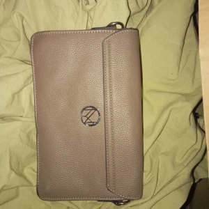 Beige axelväska från NYPD - Säljer en snygg beige axelväska från NYPD. Väskan är i skinn och har en stilren design med ett metallspänne framtill. Insidan är mönstrad och har en praktisk dragkedja. Perfekt för både vardag och fest!