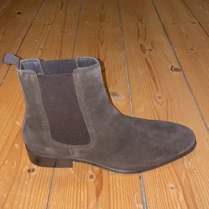 Bruna mocka Chelsea boots - Snygga Chelsea boots i brun mocka, perfekta för höst och vår. De har en klassisk design med elastiska paneler på sidorna för enkel på- och avtagning. Skorna har en låg klack och en spetsig tå som ger en stilren look. Passar både till vardags och lite mer uppklädda tillfällen. Defekt på sista sliden, därav pris