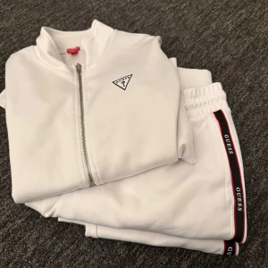 Vit hoodie från Guess - Säljer en stilren vit hoodie från Guess med dragkedja och logotyp på bröstet. Perfekt för en avslappnad look. Den har en bekväm passform och är tillverkad i mjukt material.