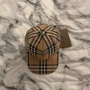 Burberry keps - Snygg rutig keps från Burberry i klassiskt beige, svart och rött mönster. Kepsen har justerbar passform och är perfekt för en stilren look. Märkeslogga på baksidan ger en exklusiv touch.