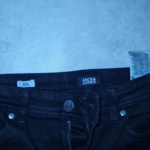 Svarta jeans från Jack & Jones - Snygga svarta jeans från Jack & Jones med slim passform. De har en klassisk design med knappar och dragkedja framtill. Perfekta för en stilren look.