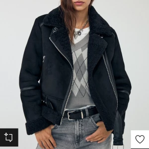 Svart jacka med fuskpäls - Snygg svart jacka med fuskpäls och dragkedja. Jackan har en klassisk bikerstil med bälte i midjan och detaljer i skinn. Perfekt för kyliga dagar.