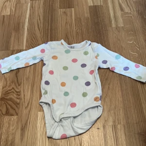 Vit prickig body från H&M - Söt vit body från H&M i ekologisk bomull med färgglada prickar i rosa, grönt, lila och orange. Den har långa ärmar och praktiska knappar i grenen för enkel på- och avklädning. Perfekt för de små! 🌈