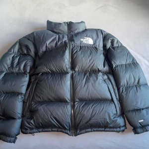 Svart dunjacka från The North Face - Säljer en svart dunjacka från The North Face med dragkedja och hög krage. Jackan har en pufferdesign och är perfekt för kalla dagar. Den har långa ärmar och logotypen syns tydligt på bröstet.