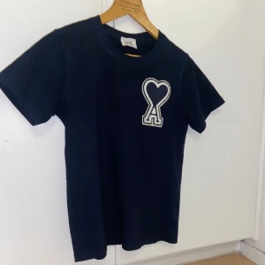 Mörkblå t-shirt från Ami - Mörkblå svart t-shirt från Ami med ett coolt hjärta och bokstaven 'A' broderat på bröstet. Perfekt för en avslappnad stil, gjord i mjuk bomull. Passar både till vardags och festliga tillfällen. Ett måste i garderoben!