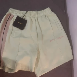 Ljus gröna shorts från Palm Angels - Säljer ett par snygga beige shorts från Palm Angels. De har en elastisk midja och coola rosa och svarta ränder på sidan. Perfekta för sommaren och avslappnade dagar. Märkesloggan är diskret broderad på framsidan. Passar perfekt för en stilren och bekväm look!