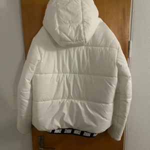 Vit pufferjacka från Nike - Säljer en snygg vit pufferjacka från Nike i mycket bra skick. Jackan har en huva med snörning och en dragkedja framtill. Perfekt för kalla dagar och har en stilren design med Nike-loggan på bröstet. Passar både till vardags och sportiga aktiviteter.