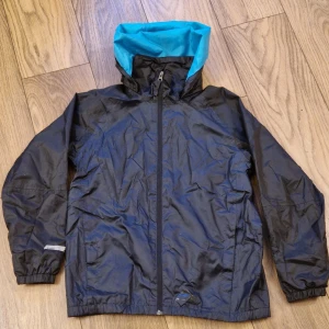Svart och blå regnjacka från McKinley - Säljer en praktisk svart regnjacka från McKinley med blå insida på huvan. Jackan har dragkedja framtill och justerbara ärmslut med kardborreband. Perfekt för regniga dagar och utomhusaktiviteter.
