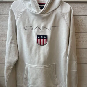 Vit hoodie från GANT - Säljer en snygg vit hoodie från GANT med broderad logga och ett coolt emblem på framsidan. Den har en klassisk känguruficka och är perfekt för både vardag och chill. Passar ungdomar i storlek 158/164 cm. Superbekväm och stilren!