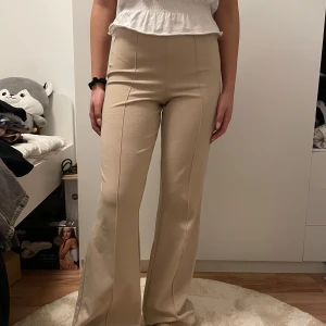 Beige byxor med utsvängda ben - Säljer ett par snygga beige byxor med hög midja och utsvängda ben. De är perfekta för både vardag och fest. Byxorna har en tight passform upptill och blir bredare nedtill, vilket ger en cool retro-look. Materialet är mjukt och bekvämt, vilket gör dem sköna att bära hela dagen.