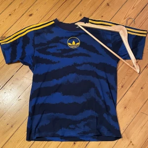 Blå och svart t-shirt från Adidas - Säljer en cool blå och svart t-shirt från Adidas med ett unikt mönster och gula detaljer. Den har korta ärmar och de klassiska tre ränderna i gult på axlarna. Perfekt för en avslappnad stil eller träning. 🏋️‍♂️