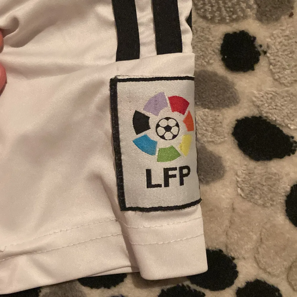 Säljer en vit Real Madrid fotbollströja från Adidas med korta ärmar. Tröjan har klubbens emblem på bröstet och LFP-logga på ärmen. Den har även 'Ronaldo 7' tryckt på ryggen. Perfekt för fans av laget! Och lite sönder de bra pris möts bara i Eskilstuna . T-shirts.