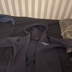 Mörkblå hoodie från Peak Performance - Säljer en mörkblå hoodie från Peak Performance med dragkedja och huva. Perfekt för kyliga dagar. Märkeslogga på bröstet ger en stilren look. Passar både till vardags och avslappnade tillfällen. Size: L för barn