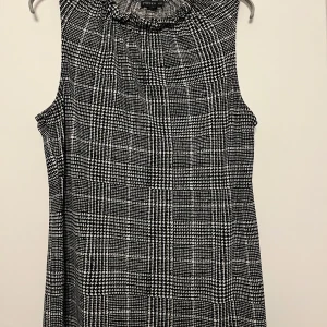 Svartvit ärmlös top/blus från Stockh LM - Svartvit ärmlös top/blus från Stockh LM (MQ) med ett klassiskt rutigt mönster. Toppen/blusen har en hög krage med volangdetaljer. Fin under en kofta eller kavaj.