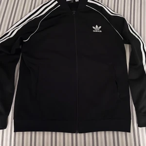 Svart träningsjacka från Adidas - Snygg svart träningsjacka från Adidas med de klassiska tre vita ränderna längs ärmarna. Jackan har dragkedja framtill och Adidas-logga på bröstet. Perfekt för en sportig look. Storlek 160 för 13 till 14 åringar