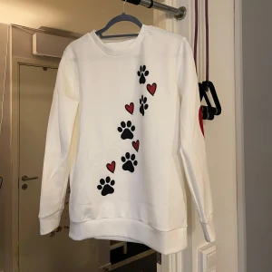 Vit sweatshirt med tassavtryck och hjärtan - Säljer en vit sweatshirt med ett gulligt tryck av svarta tassavtryck och röda hjärtan. Tröjan har långa ärmar och en rund halsringning. Perfekt för djurälskare! 🐾❤️helt ny