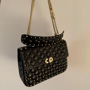 Svart quiltad axelväska med nitar - Snygg svart axelväska i skinn med quiltad design och coola nitar. Den har en elegant kedjerem och ett praktiskt spänne framtill. Insidan är rymlig med en röd ficka med dragkedja. Perfekt för både vardag och fest!