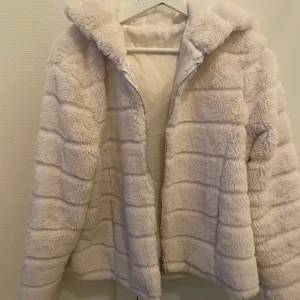 Vit teddyjacka i polyester - Mysig vit teddyjacka i 100% polyester. Jackan har en dragkedja framtill och en stor huva för extra värme. Det är dock hål i fickorna.