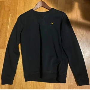 Svart tröja från Lyle & Scott - Säljer en stilren svart tröja från Lyle & Scott med deras ikoniska gula logga på bröstet. Tröjan har långa ärmar och en klassisk rund halsringning. Perfekt för både vardag och lite finare tillfällen. Passar bra till jeans eller chinos!