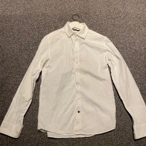 Vit skjorta från Tiger of Sweden - Säljer en stilren vit skjorta från Tiger of Sweden i slim fit. Skjortan är långärmad och har klassiska knappar framtill. Perfekt för både formella och avslappnade tillfällen. Materialet känns mjukt och bekvämt. Passar utmärkt till både kostym och jeans!