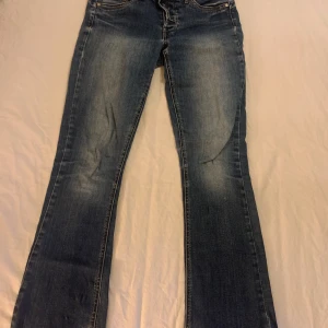 Blå jeans med bootcut - Säljer ett par snygga blå jeans i bootcut-stil. De har en cool slitningseffekt och är i bra skick. Perfekta för en avslappnad look. Detaljer som knappar med ett unikt mönster ger dem en extra touch. Passar perfekt till både vardag och fest!