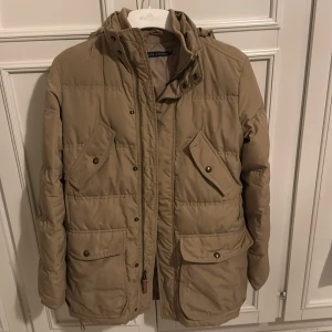 Beige dunjacka från Ralph Lauren - Säljer en snygg beige dunjacka från Ralph Lauren i storlek XS. Jackan är i mycket bra skick och perfekt för höst och vinter. Den har långa ärmar och praktiska fickor med knappar. Materialet är polyester, vilket gör den både varm och lätt. Perfekt för att hålla stilen när det blir kallt ute!