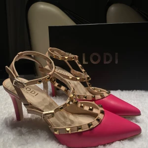 Rosa pumps med nitar från Lodi - Säljer ett par snygga rosa pumps från Lodi med spetsig tå och klack. De har coola guldfärgade nitar som ger en edgy look. Perfekta för fest eller en utekväll! Använda endast en gång, så de är i mycket bra skick.