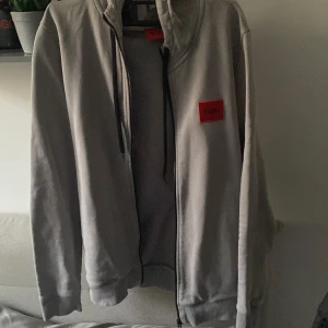 Grå hoodie från Hugo Boss - Säljer en snygg grå hoodie från Hugo Boss i mycket bra skick. Den har en dragkedja framtill och en röd logga på bröstet. Perfekt för höst och vår, med långa ärmar och en skön passform. Perfekt för en casual look eller att mysa i hemma.