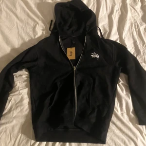 Svart hoodie från Stüssy - Säljer en svart hoodie från Stüssy med dragkedja och vit logga på bröstet och ryggen. Perfekt för en avslappnad stil med justerbar huva och långa ärmar. Passar bra till både vardag och chill.