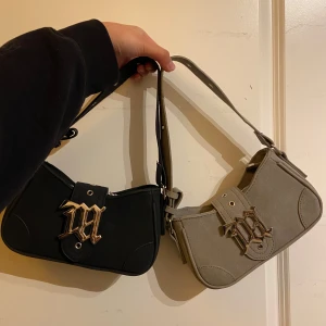 Svart och beige axelväska med metallinitial - Två snygga axelväskor i svart och beige med en cool metallinitial på framsidan. Väskorna har en dragkedja upptill och justerbara axelremmar. Perfekt för att ge din outfit en edgy touch! 🖤