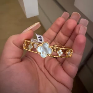 Guldarmband med klöver - Snyggt guldarmband med  klöver och vita detaljer. Armbandet har en elegant design med flera blommor i pärlemor och gnistrande stenar. Perfekt för att ge en touch av glamour till vilken outfit som helst. Passar både till vardags och festliga tillfällen ny oanvänd .