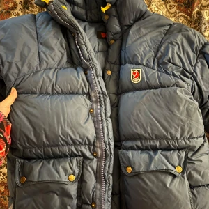 Blå dunjacka från Fjällräven - Säljer en snygg blå dunjacka från Fjällräven i mycket bra skick. Jackan har en hög krage och praktiska fickor med knappar. Perfekt för kalla vinterdagar. Den har en dragkedja och knappar framtill för extra skydd mot kylan.