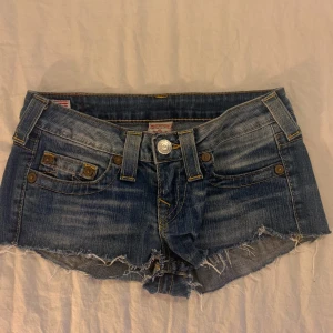 Blå jeansshorts från True Religion - Säljer ett par snygga blå jeansshorts från True Religion i bra skick. De är lågmidjade med slitna kanter och klassiska femficksdesign. Perfekta för sommardagar eller festivaler! 🕶️