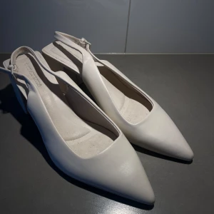 Vita pumps med klack - Eleganta vita pumps med spetsig tå och bred klack. Skorna har en stilren design med en justerbar rem runt hälen för bättre passform.