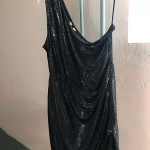 Svart glittrig klänning - Snygg svart glittrig klänning med en axelbandslös design. Perfekt för en kväll ute med vänner. Klänningen har en draperad stil som ger en elegant look.