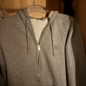 Grå hoodie från Burberry - Säljer en stilren grå hoodie från Burberry med dragkedja och broderad logga på bröstet. Den har en klassisk huva med dragsko och är tillverkad i mjukt material för extra komfort. Perfekt för en avslappnad look. Det är en storlek M. 