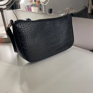 Svart axelväska med krokodilmönster - Snygg svart axelväska i syntet med krokodilmönster. Väskan har en dragkedja upptill. Perfekt för att ge en stilren touch till din outfit.